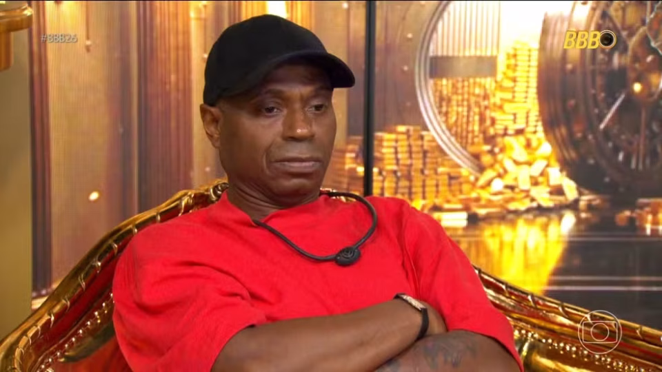 Frame de vídeo mostra Edilson, que usa camiseta vermelha e boné preto, sentado, de braços cruzados, no confessionário do BBB 26